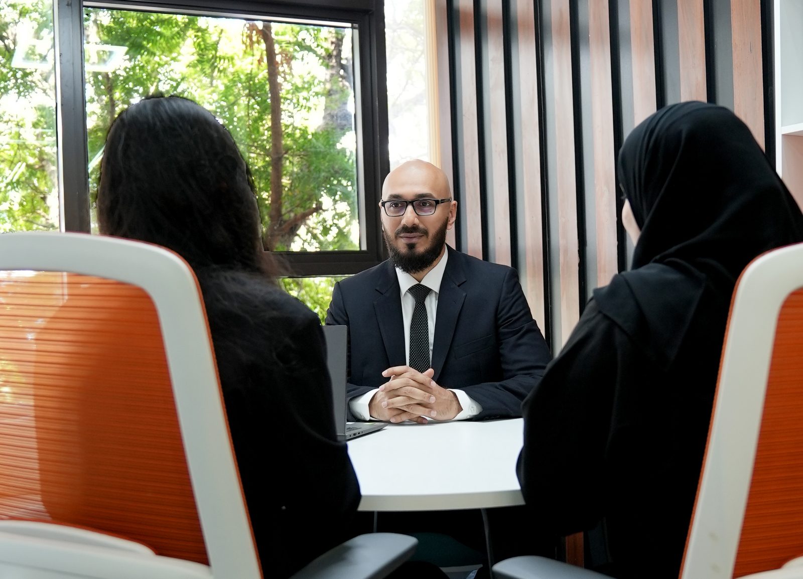 An advisor in a navy suit facing two clients across a white table, with a window and greenery behind him.