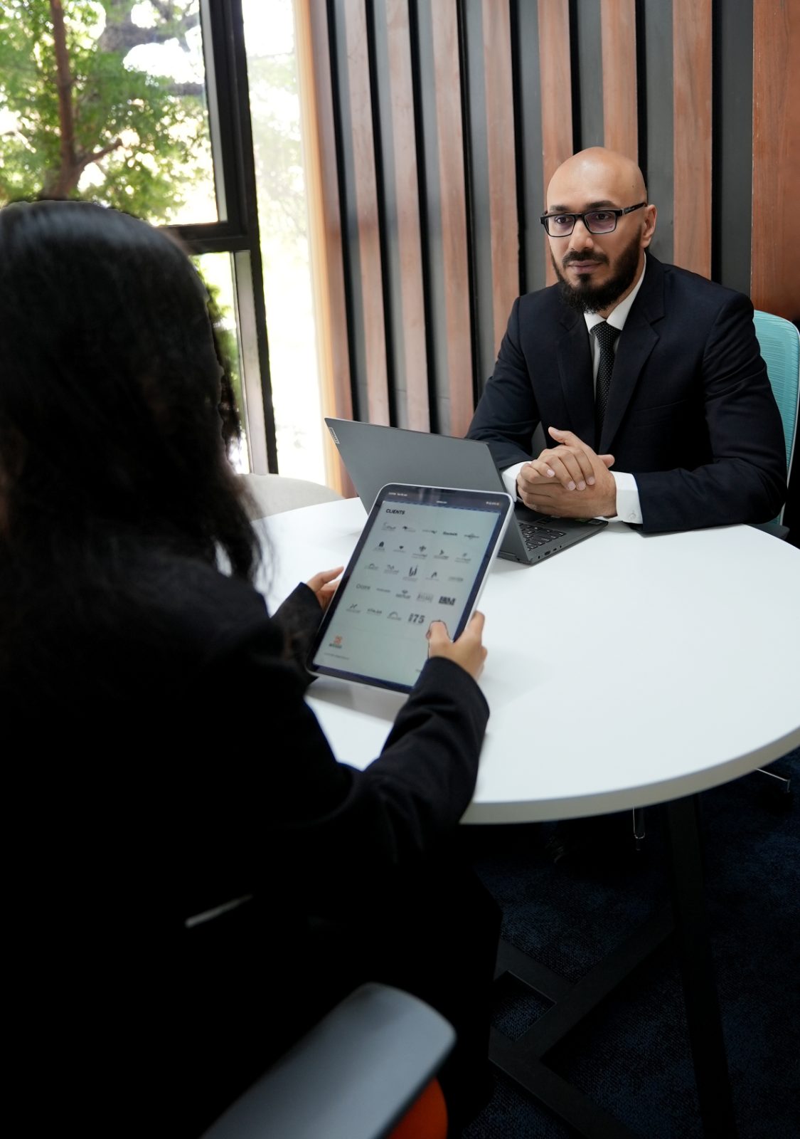 A client holding a tablet showing the Sella clients page, with an advisor in a dark suit seated opposite at a round white table.